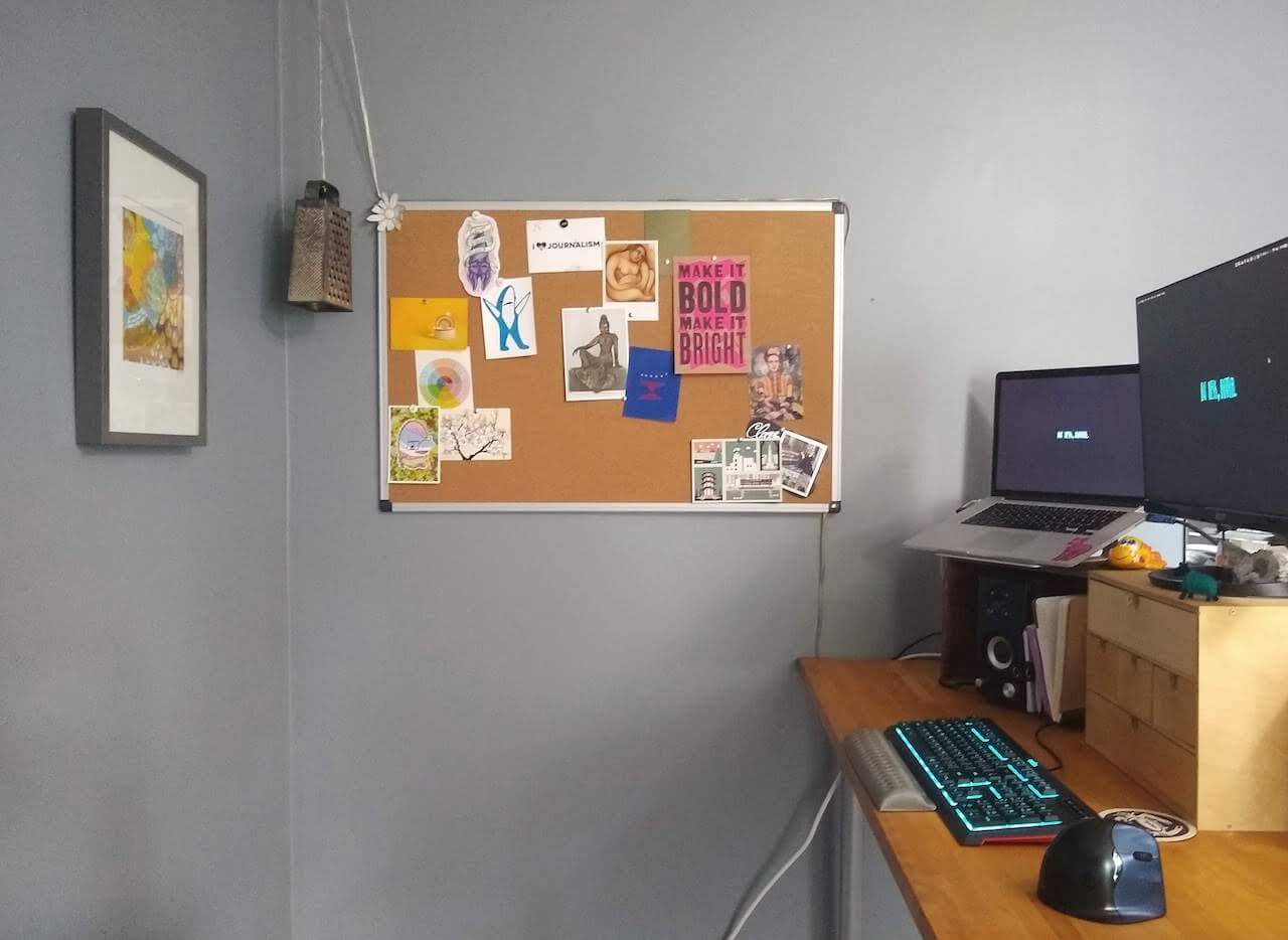 My standing desk with its accoutrements: a MacBook and peripherals, like an external monitor, keyboard, vertical mouse and speakers. The walls are painted a soothing blue grey and are decorated with artwork and a corkboard, also covered in art and postcards.